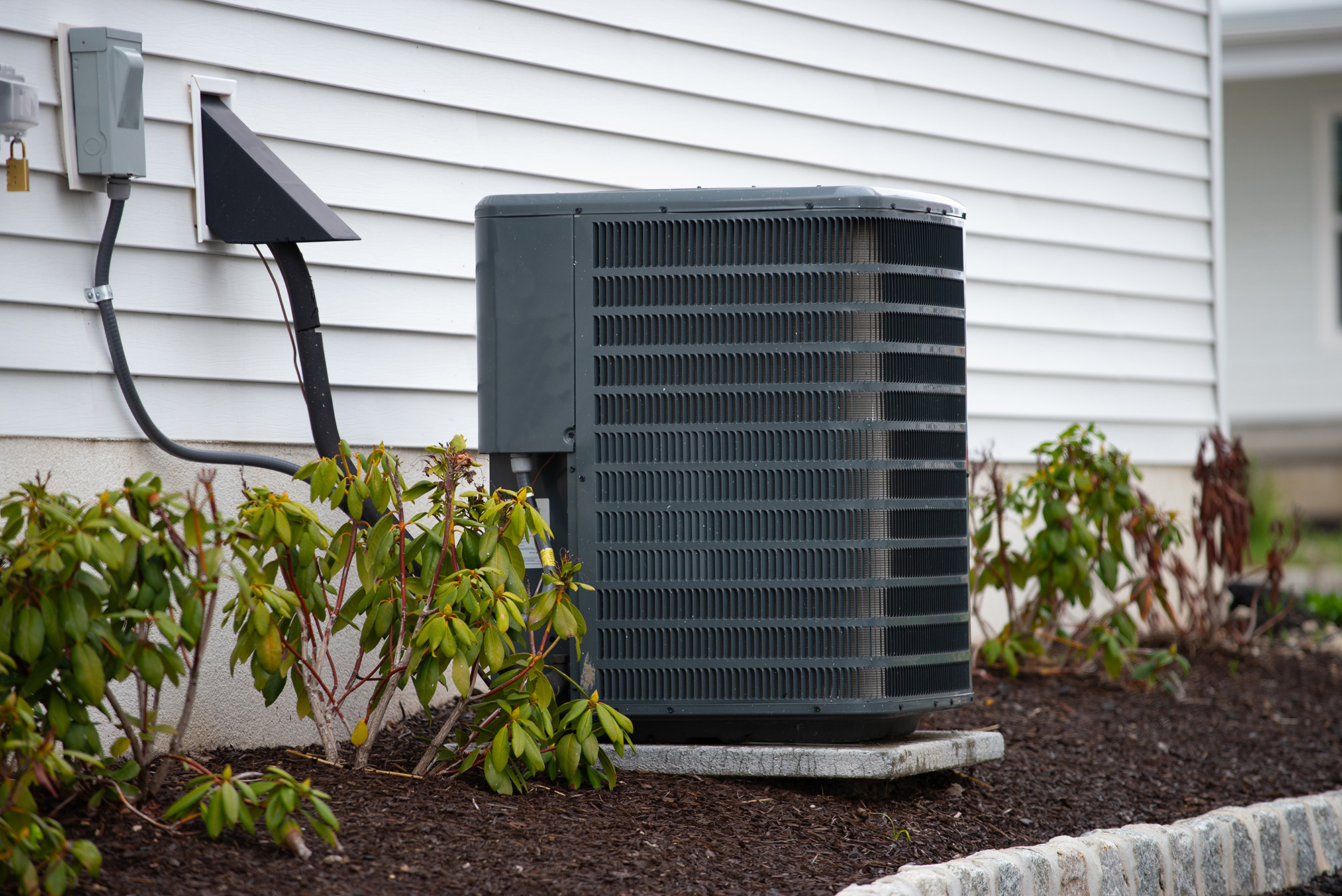 A large outdoor heat pump sits on a concrete pad next to a white house, surrounded by mulch and small green shrubs.
