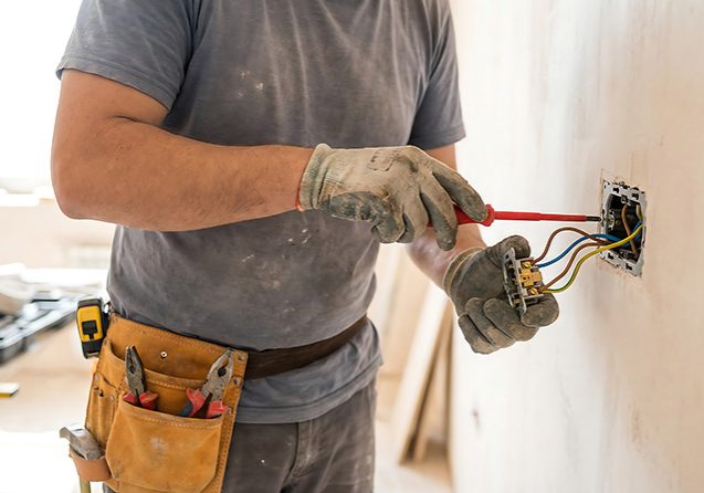 A person wearing gloves and a tool belt is installing or repairing electrical wiring in a wall, using a screwdriver. Various colored wires are visible, and tools are scattered on a surface in the background.