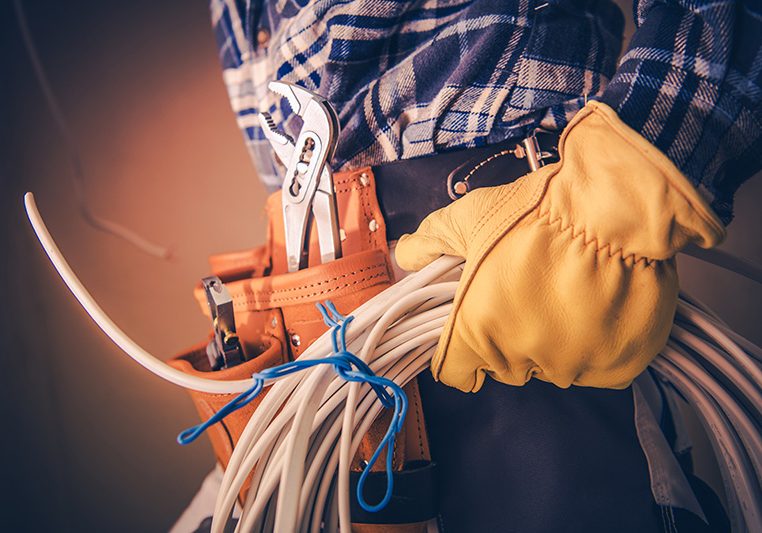 Person wearing a tool belt and yellow work gloves holds a bundle of electrical cables. Tools like pliers and screwdrivers are visible in the belt. The person is dressed in a blue plaid shirt.
